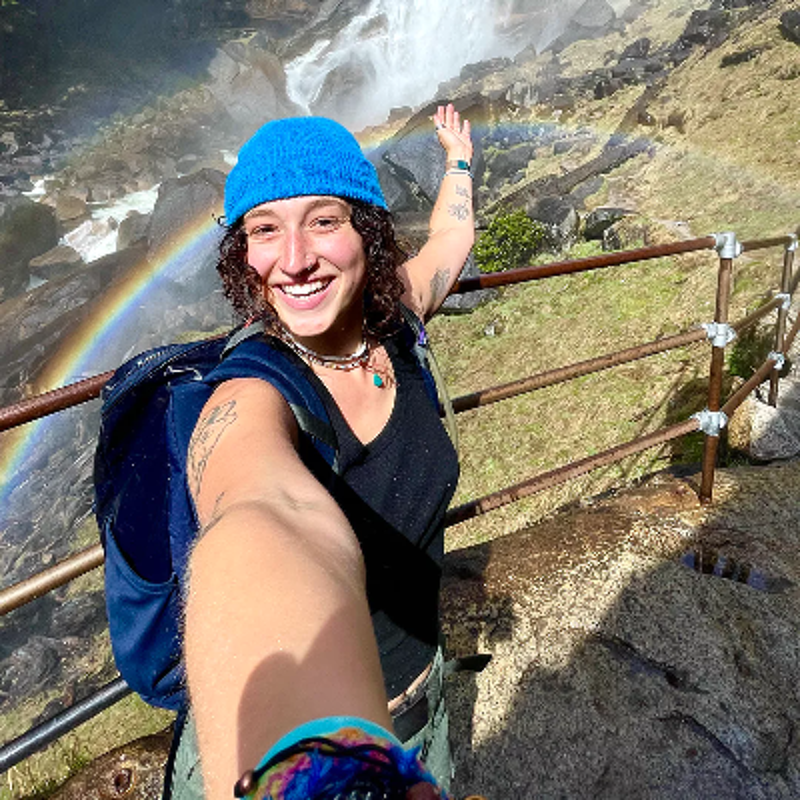 The image shows a person smiling widely and taking a selfie. They are wearing a blue hat, a black shirt, and have a backpack. A rainbow arcs across the scene, and a waterfall is visible in the background. The person is standing on a path with a railing.